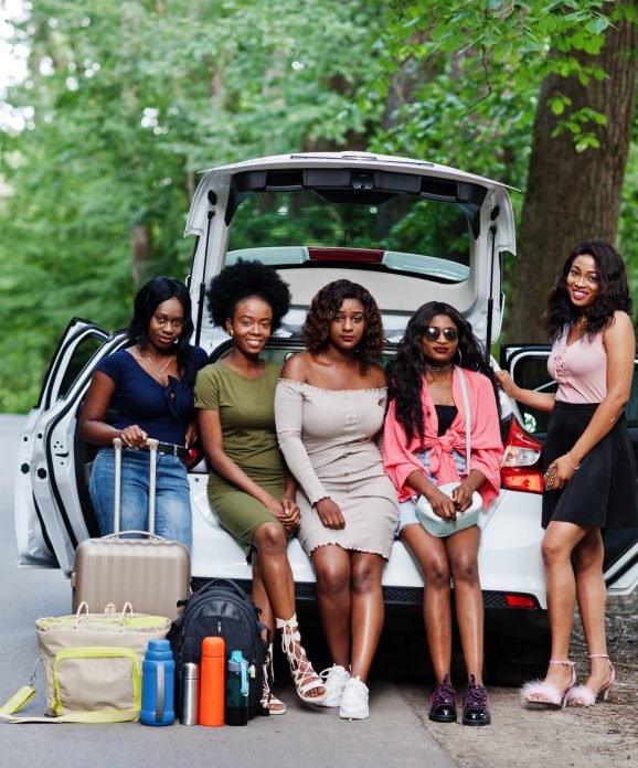 Group of five happy african american traveler girls sitting in car open trunk.